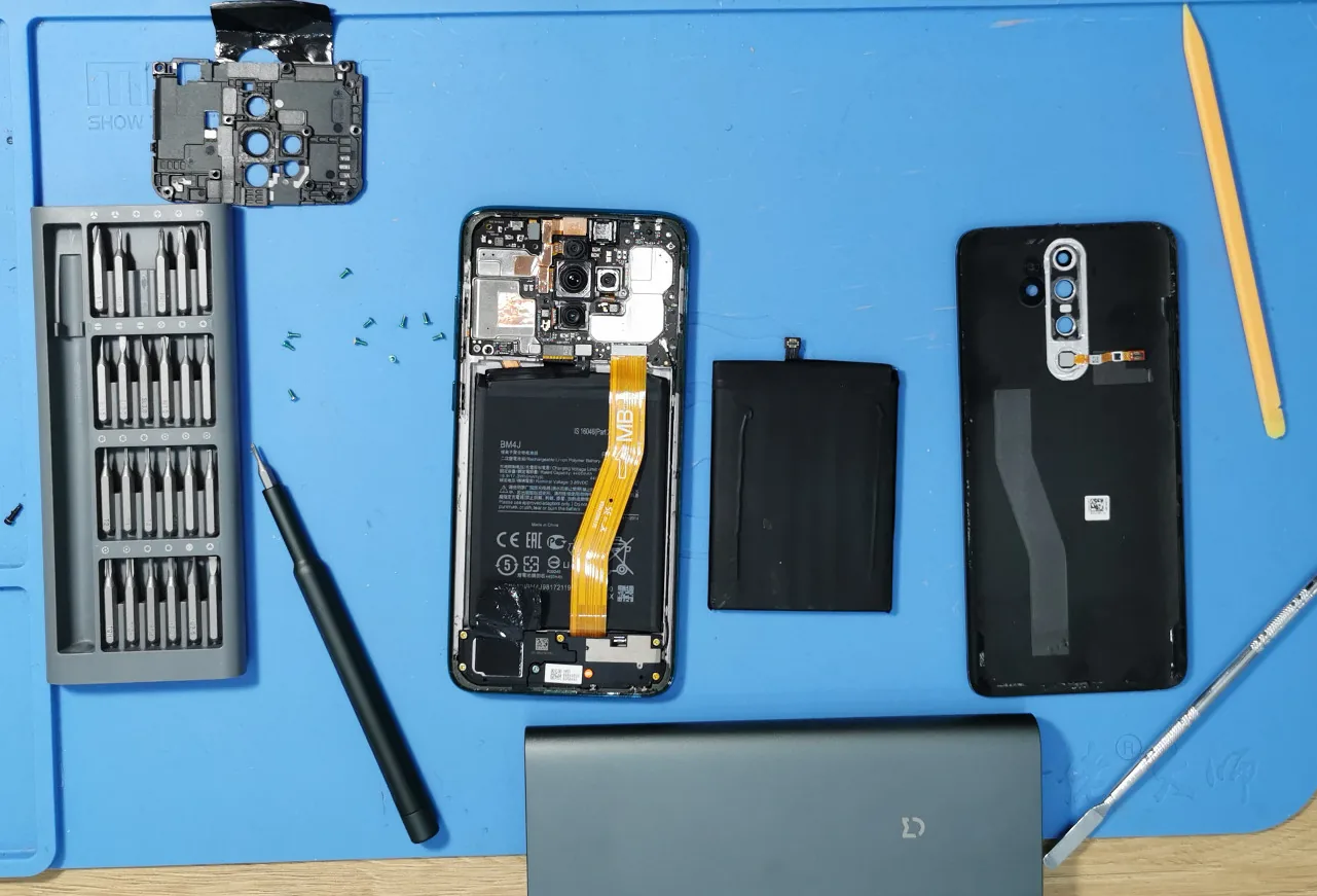 Синий силиконовый коврик мастера: вскрытый Xiaomi без задней крышки, снятый аккумулятор, набор бит, лопатка и пинцет, рядом пауэрбанк.