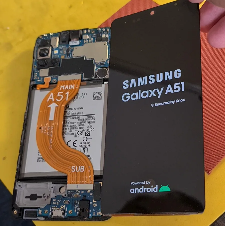 Разобранный Samsung Galaxy A51 на жёлтом столе мастерской: слева корпус с платой, аккумулятором и оранжевым шлейфом, справа снятый дисплей с логотипом Samsung.