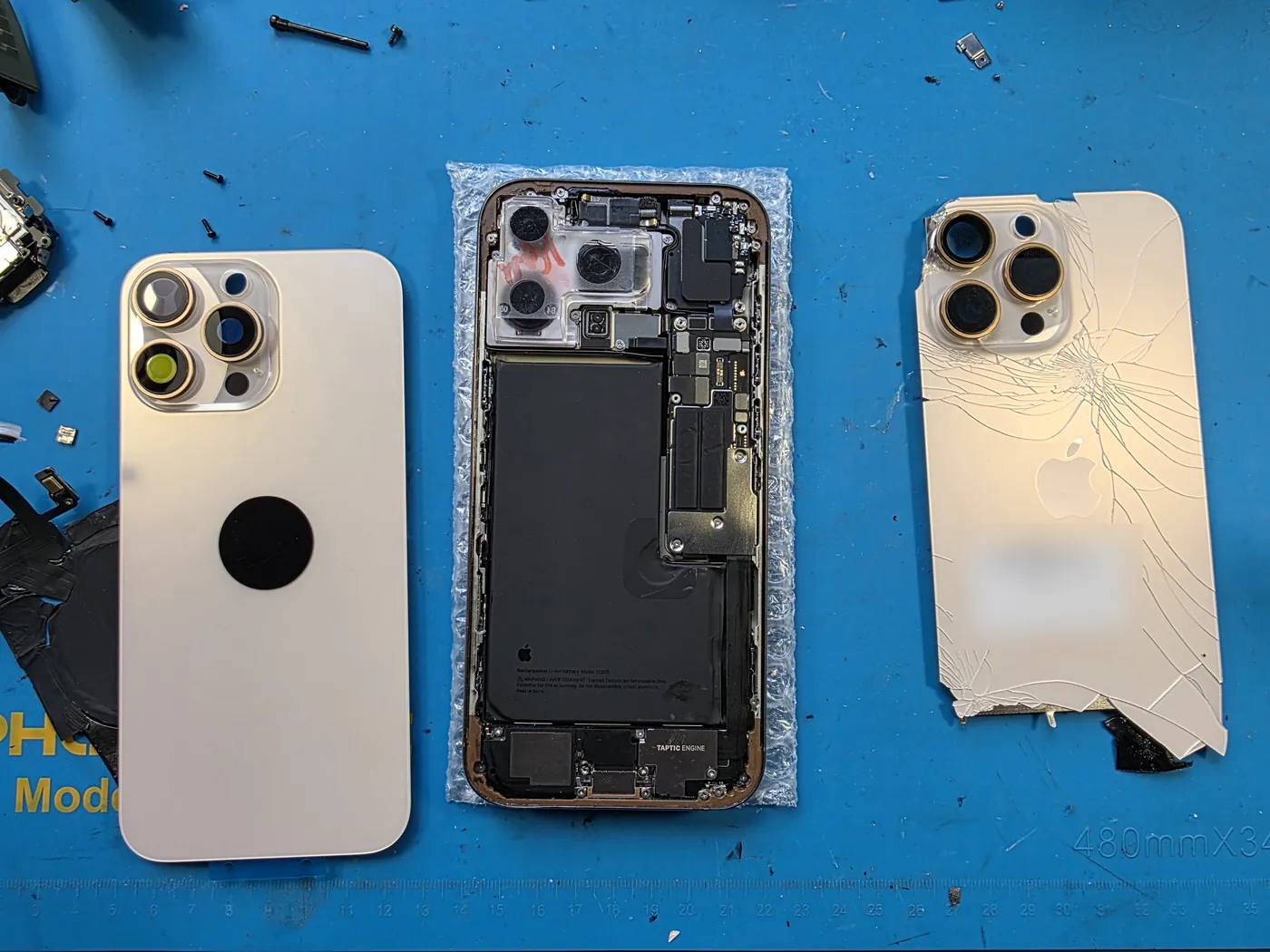 Разобранный iPhone 16 Pro Max на синем коврике сервиса: посередине корпус с платой и аккумулятором, слева новая задняя крышка, справа снятая, вся в трещинах.