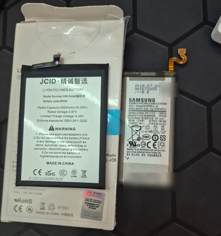 Два аккумулятора на столе: новый JCID для Redmi Note 9 на своей коробке и снятый аккумулятор Samsung с датой 2018 года на наклейке.