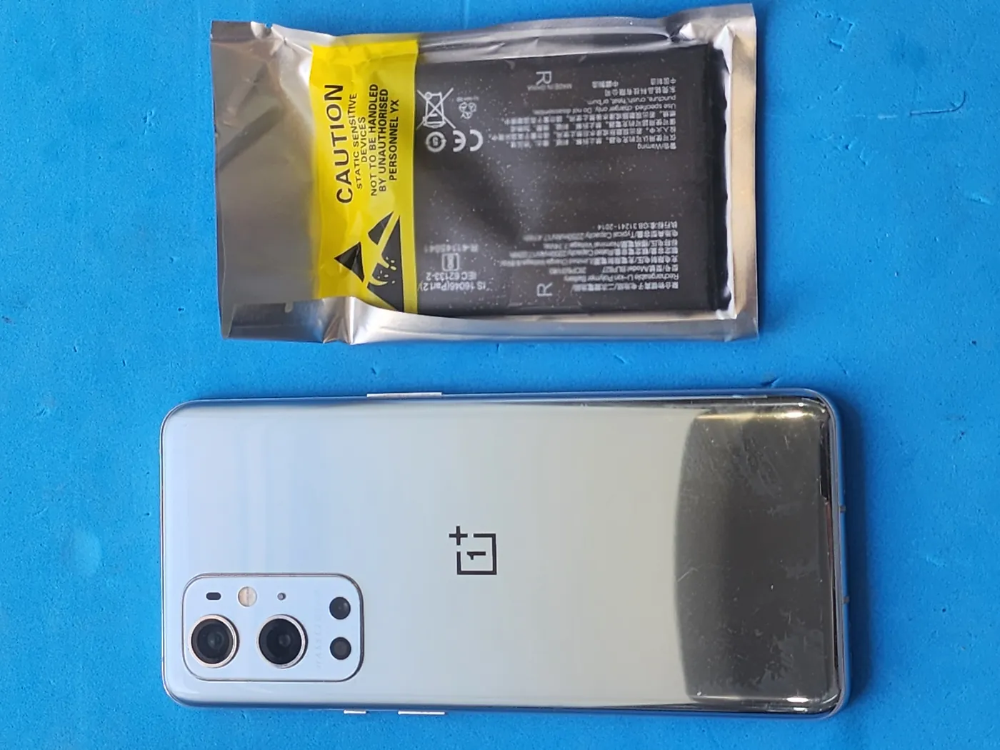 Смартфон OnePlus задней крышкой вверх на синем коврике, над ним новый аккумулятор в антистатическом пакете с жёлтой наклейкой.